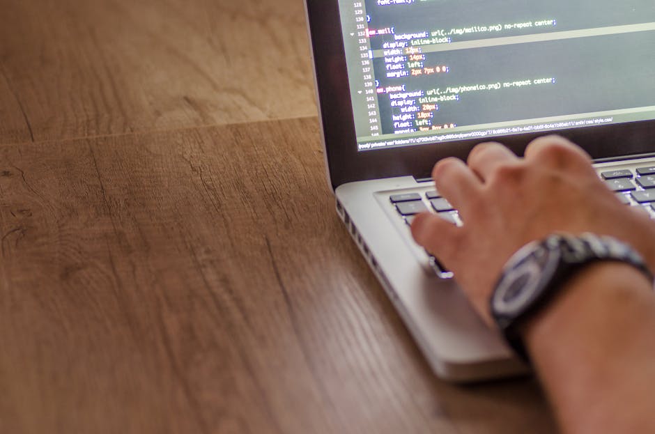 A person using a laptop placed on a wooden desk, with a focus on their right hand positioned over the keyboard. The laptop screen displays lines of code in a text editor. The person's wrist is adorned