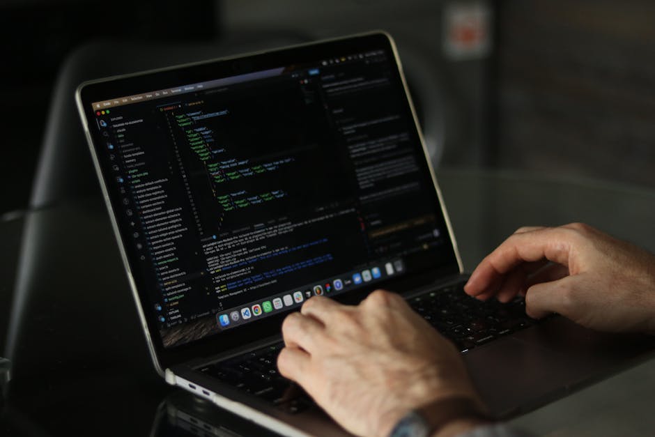 A person using a laptop placed on a wooden desk, with a focus on their right hand positioned over the keyboard. The laptop screen displays lines of code in a text editor. The person's wrist is adorned