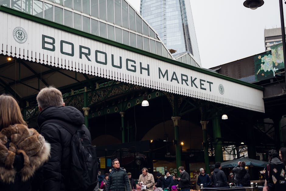 A lively outdoor market scene with a bakery stall featuring a large stack of freshly baked bread and loaves displayed behind a glass front. Two elderly men, one wearing a black beanie and the other a 